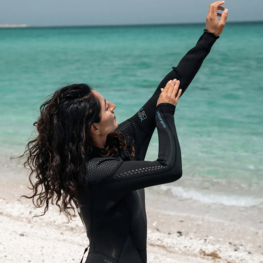 Woman in a Fourth Element Xenos ARC Wetsuit stretching on a beach with clear blue water in the background